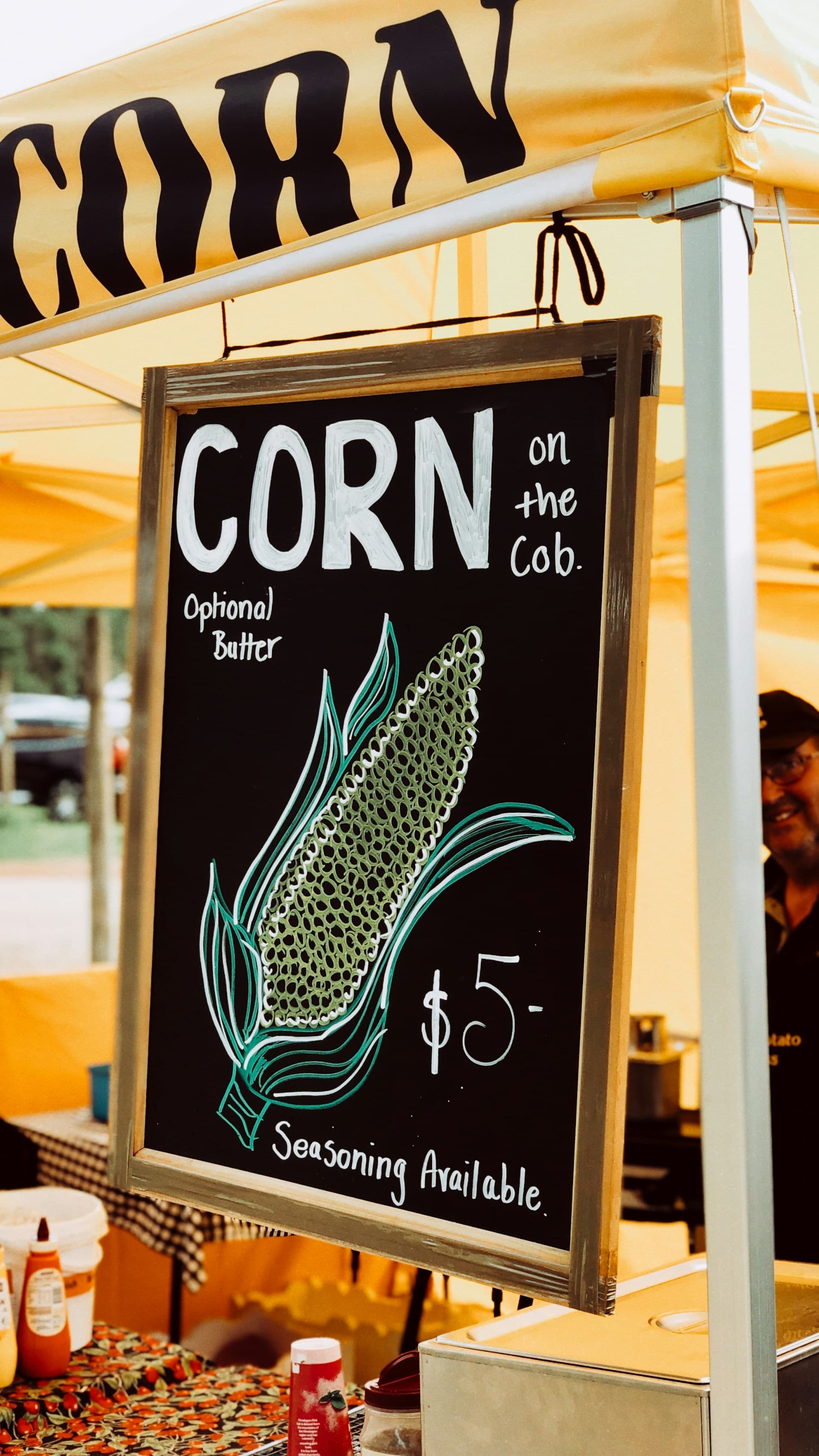 A hand-lettered farm-stand A-frame reading 'CORN on the cob' with a chalk illustration of corn.