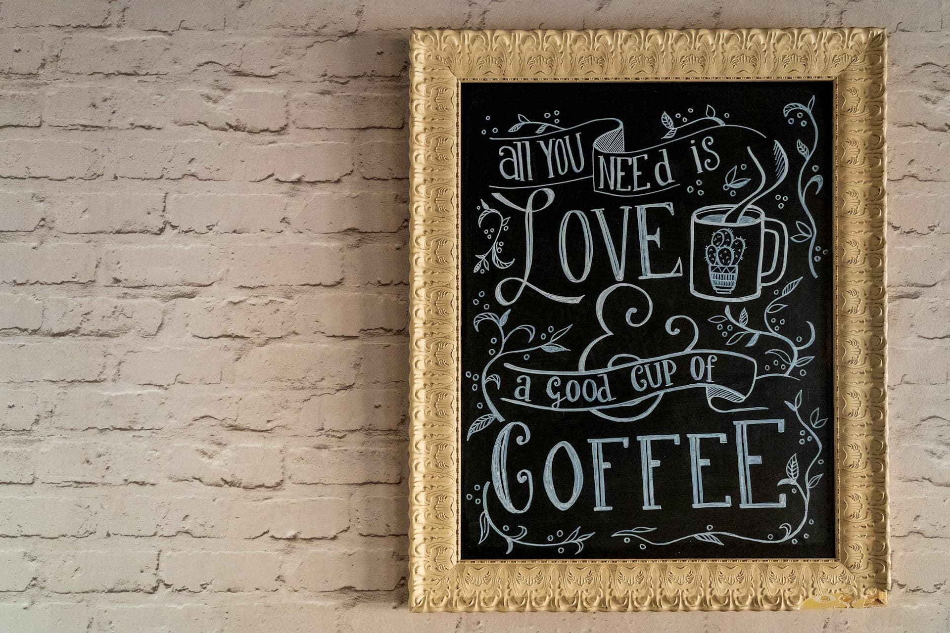 Framed chalk piece reading 'All you need is love & a good cup of coffee' on a white brick wall.